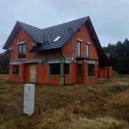 Surowy stan domu z czerwonej cegły z ciemną dachówką, widoczne okna i drzwi zabite deskami, na pierwszym planie skrzynka elektryczna, pochmurne niebo.