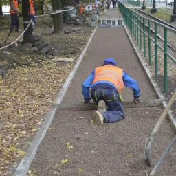 Pracownik w odblaskowej kamizelce wyrównuje podłoże pod ścieżkę z drobnego kruszywa wzdłuż torów tramwajowych. Widoczne narzędzia i krawężniki.