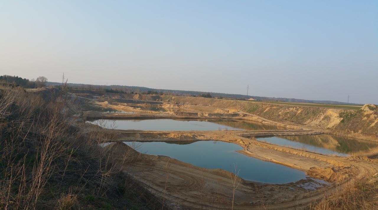 Kopalnia kruszywa z widocznymi zbiornikami wodnymi, śladami opon i brzegami z piasku oraz ziemi, w tle las i pole uprawne pod błękitnym niebem.