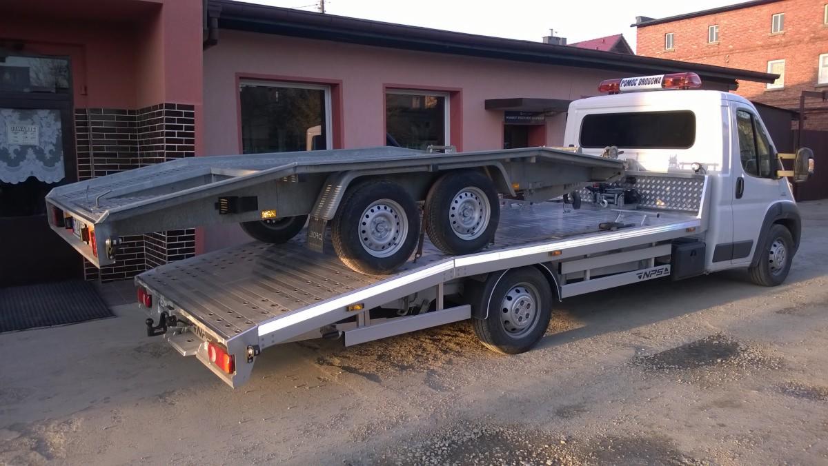 Laweta z przyczepą na samochodzie pomocy drogowej, gotowa do transportu. Widoczny budynek w tle. Pomoc drogowa z sygnalizacją świetlną na dachu.