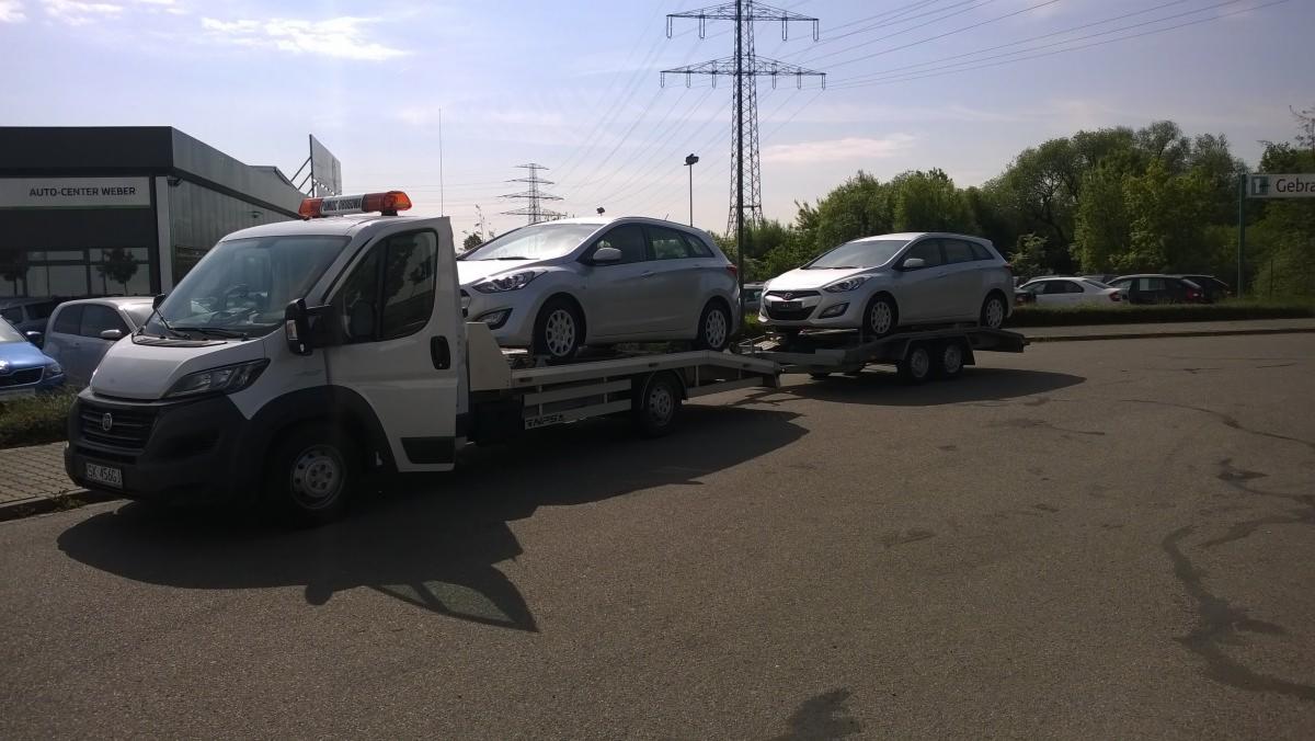 Laweta z dwoma srebrnymi autami Hyundai i30 na parkingu, w tle słupy wysokiego napięcia i budynek Auto-Center Weber.