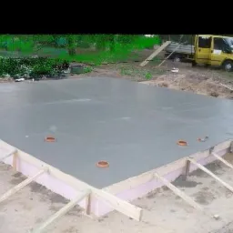 Wylana, świeża płyta fundamentowa na różowej izolacji, z widocznymi rurami kanalizacyjnymi, drewnianym szalunkiem i żółtym samochodem dostawczym w tle.