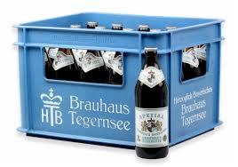Błękitna skrzynka z piwem Brauhaus Tegernsee i butelka tego piwa na pierwszym planie. Widoczne logo browaru i etykiety butelek w skrzynce.