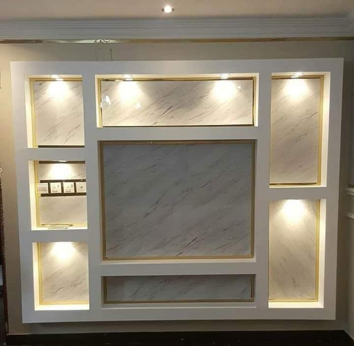 Biała ściana z wnękami wykończonymi marmurkową płytą i złotymi obramowaniami, oświetlona punktowymi lampami, z widocznymi gniazdkami elektrycznymi w jednej z wnęk.