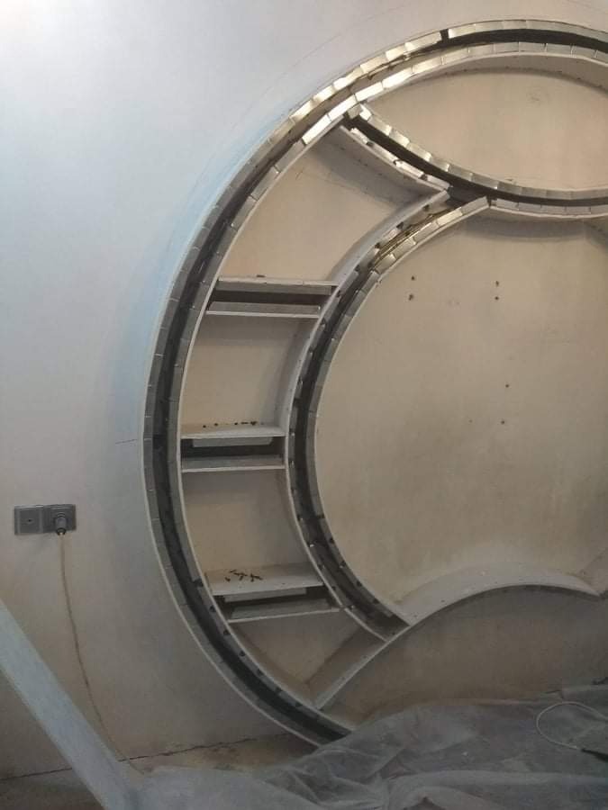 Niedokończona okrągła wnęka z półkami w ścianie, widoczne metalowe profile i fragment instalacji elektrycznej.