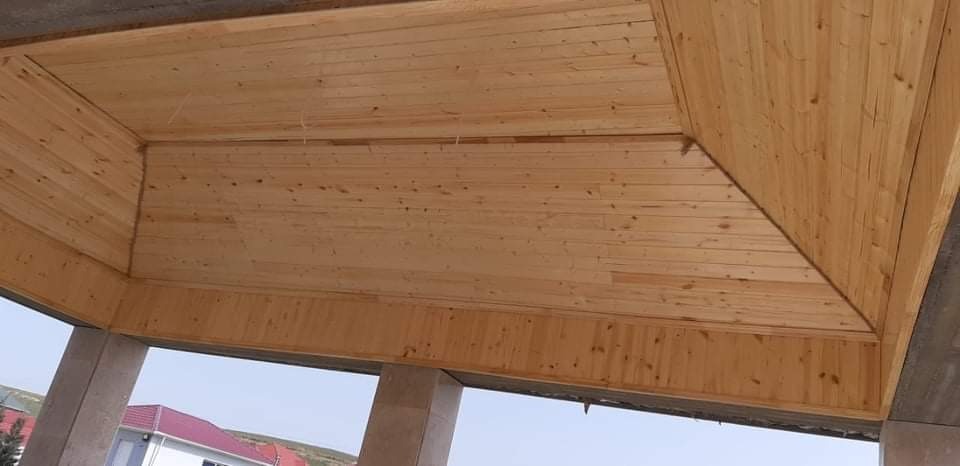 Drewniana podbitka dachowa w jasnym odcieniu, widoczna od spodu, surowe betonowe słupy w tle, fragment budynku z czerwoną dachówką w oddali.