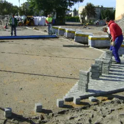 Roboty brukarskie w toku: ułożona część nawierzchni z szarej kostki, palety z kostką Libet w tle, pracownicy przy pracy, widoczny sprzęt budowlany.