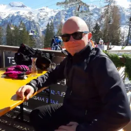 Mężczyzna w okularach przeciwsłonecznych siedzi przy żółtym stoliku na tle górskiego krajobrazu z wyciągiem narciarskim i śnieżnymi szczytami.