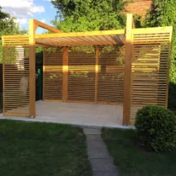 Nowoczesna altana z drewna z poziomymi listwami, wykończona na jasny odcień, postawiona na płytach chodnikowych, otoczona zielenią ogrodu.