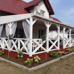 Zadaszony taras z białymi, drewnianymi balustradami w kształcie krzyży, czerwonym podestem i zwiewnymi, białymi zasłonami, otoczony świeżo posadzoną roślinnością i obrzeżem z kostki granitowej.