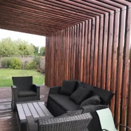 Nowoczesna konstrukcja z ciemnego drewna tworząca zadaszony taras z elementami pionowych paneli, wyposażona w czarne meble ogrodowe z technorattanu i drewniany stolik kawowy.