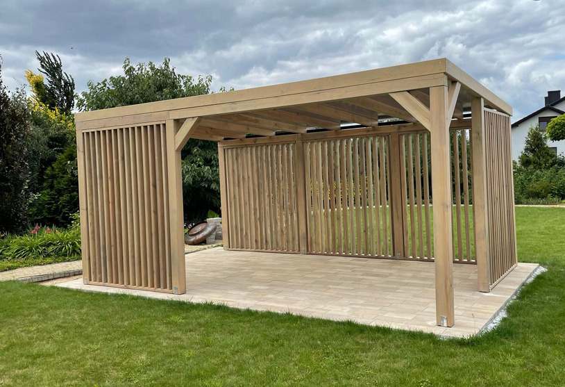 Nowoczesna, drewniana konstrukcja z pionowymi listwami, tworząca zadaszoną przestrzeń na tarasie wyłożonym kostką, otoczona zielonym trawnikiem i drzewami.