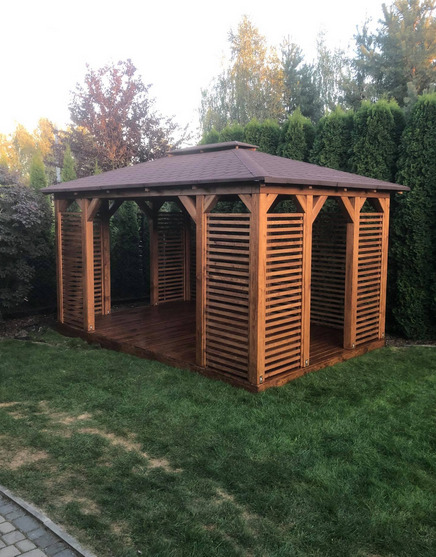 Drewniana altana z brązowym dachem i poziomymi listwami, ustawiona na trawniku w ogrodzie z tujami w tle.