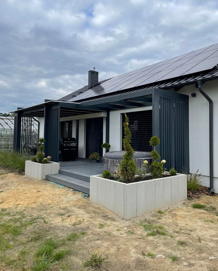 Nowoczesny taras z szarej deski kompozytowej, zadaszony pergolą w kolorze antracytowym, z dwoma betonowymi donicami z roślinami i jacuzzi w tle, widok na dom z panelami słonecznymi.