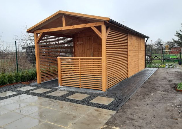 Nowoczesna drewniana konstrukcja z częścią zamkniętą i otwartą, z poziomymi listwami, na szarym brukowanym tarasie z elementami dekoracyjnymi z kamyków.