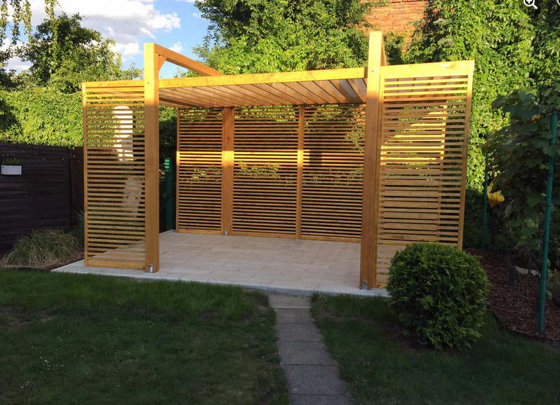 Nowoczesna altana z drewna z poziomymi listwami, wykończona na jasny odcień, postawiona na płytach chodnikowych, otoczona zielenią ogrodu.