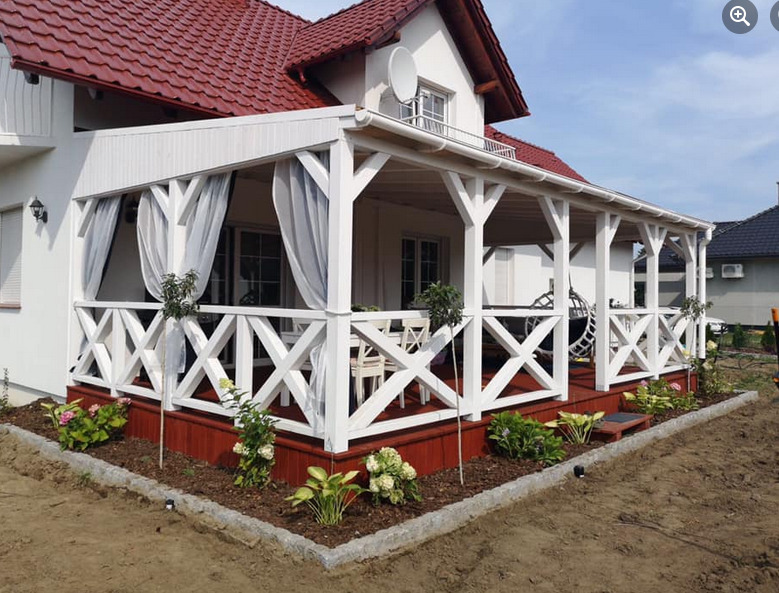 Zadaszony taras z białymi, drewnianymi balustradami w kształcie krzyży, czerwonym podestem i zwiewnymi, białymi zasłonami, otoczony świeżo posadzoną roślinnością i obrzeżem z kostki granitowej.