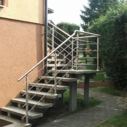 Zewnętrzne schody z poręczami ze stali nierdzewnej, prowadzące do budynku z beżową elewacją. Widok na zielony żywopłot w tle. Schody z szarego kamienia.