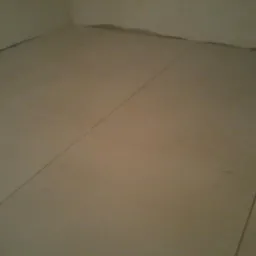 Beżowe płytki podłogowe ułożone w rogu pomieszczenia. Widoczne spoiny i niedokończone wykończenie przy ścianach. Brak listew przypodłogowych.