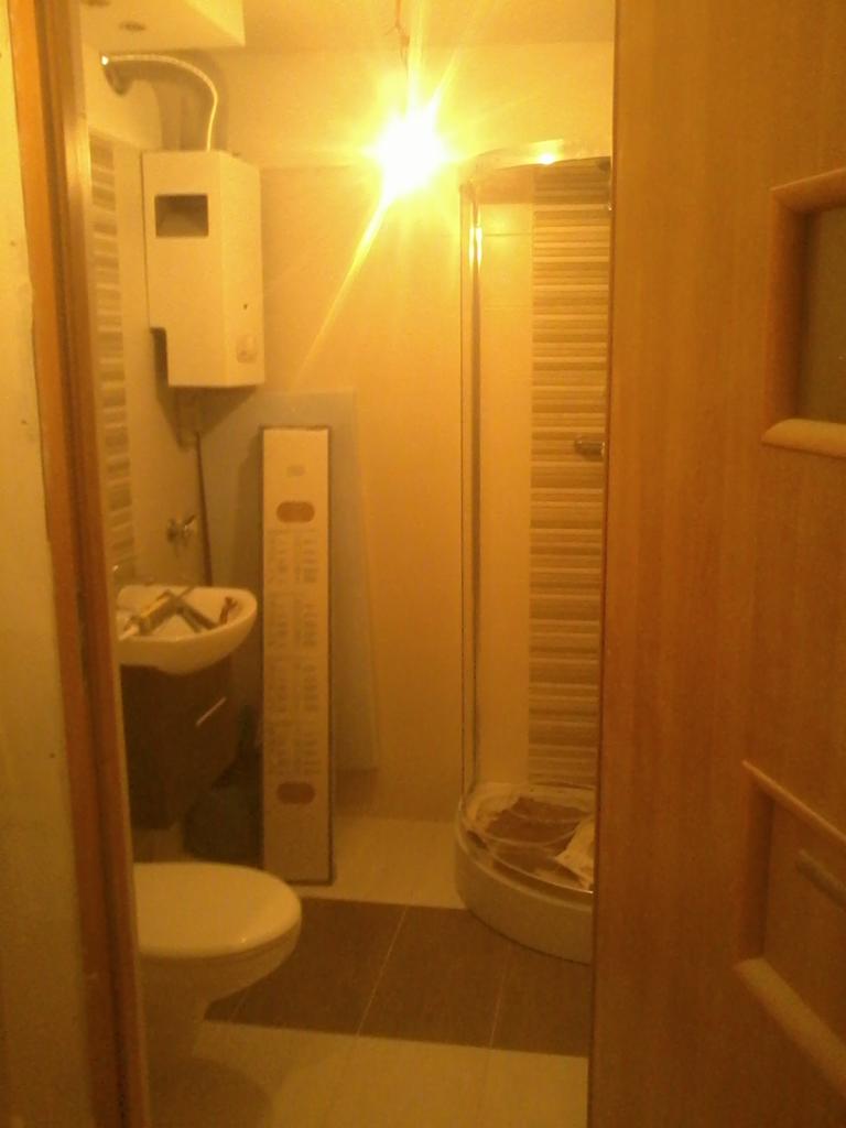 Ukończona łazienka z kabiną prysznicową, umywalką i toaletą. Widok zza uchylonych drzwi, ciepłe oświetlenie.