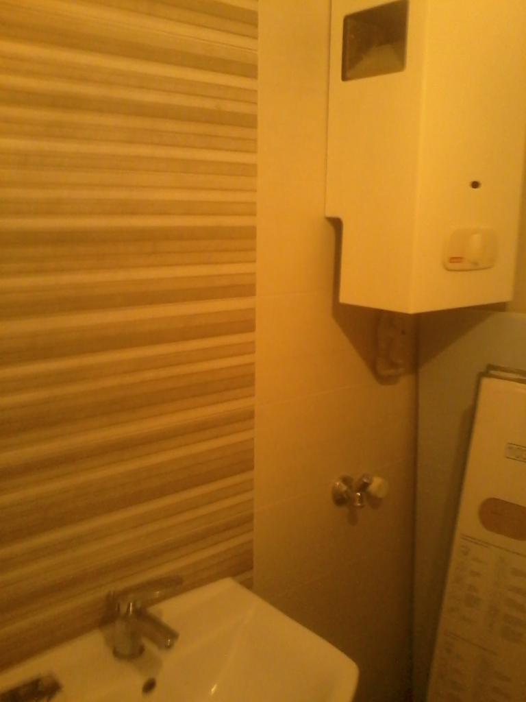 Umywalka w łazience z poziomymi, beżowymi płytkami na ścianie. Widoczny podgrzewacz wody i bateria umywalkowa. Całość oświetlona ciepłym światłem.