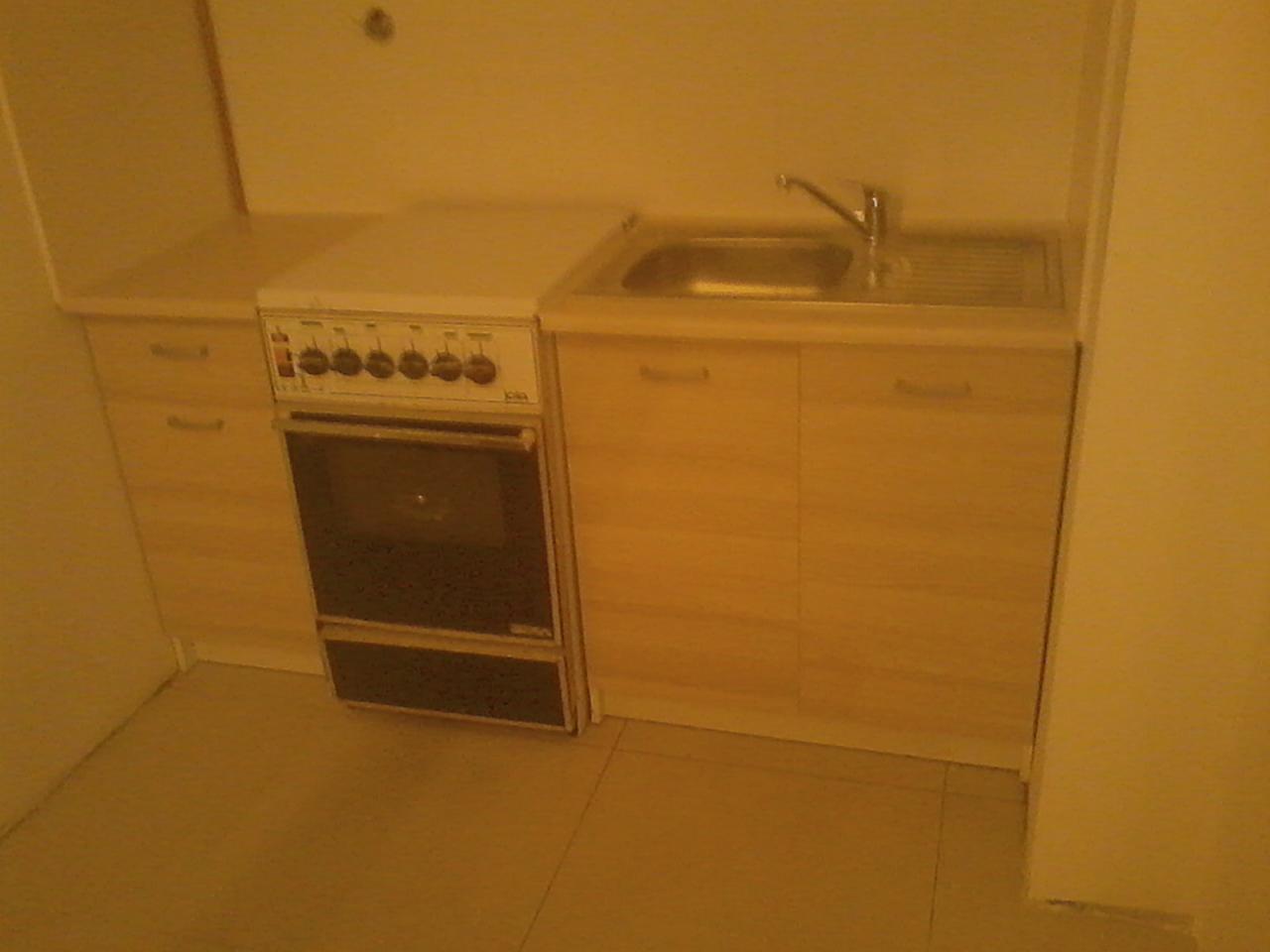 Kompaktowa kuchnia z drewnopodobnymi frontami szafek, wyposażona w zlewozmywak i kuchenkę z piekarnikiem, w ciepłej tonacji kolorystycznej.