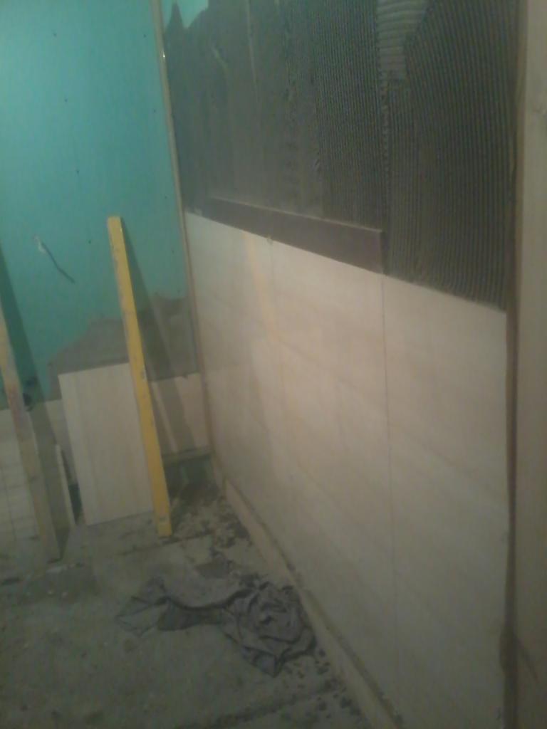 Układanie płytek ściennych w łazience, widoczny klej na ścianie, poziomica i zapas płytek. Prace glazurnicze w toku, surowe wykończenie.