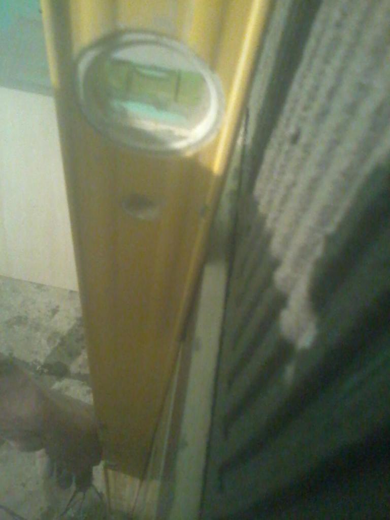 Żółta poziomica przyłożona do ściany z szarym panelem, sprawdzanie pionu. Ujęcie z dołu, niedoskonała jakość obrazu.