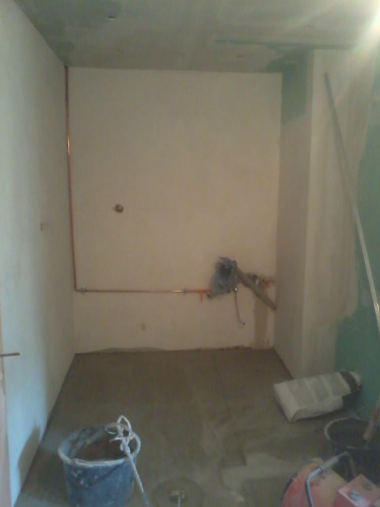 Surowy stan łazienki w trakcie prac: widoczne instalacje, przygotowane ściany, świeżo wylana posadzka jastrychowa, narzędzia i materiały budowlane na podłodze.