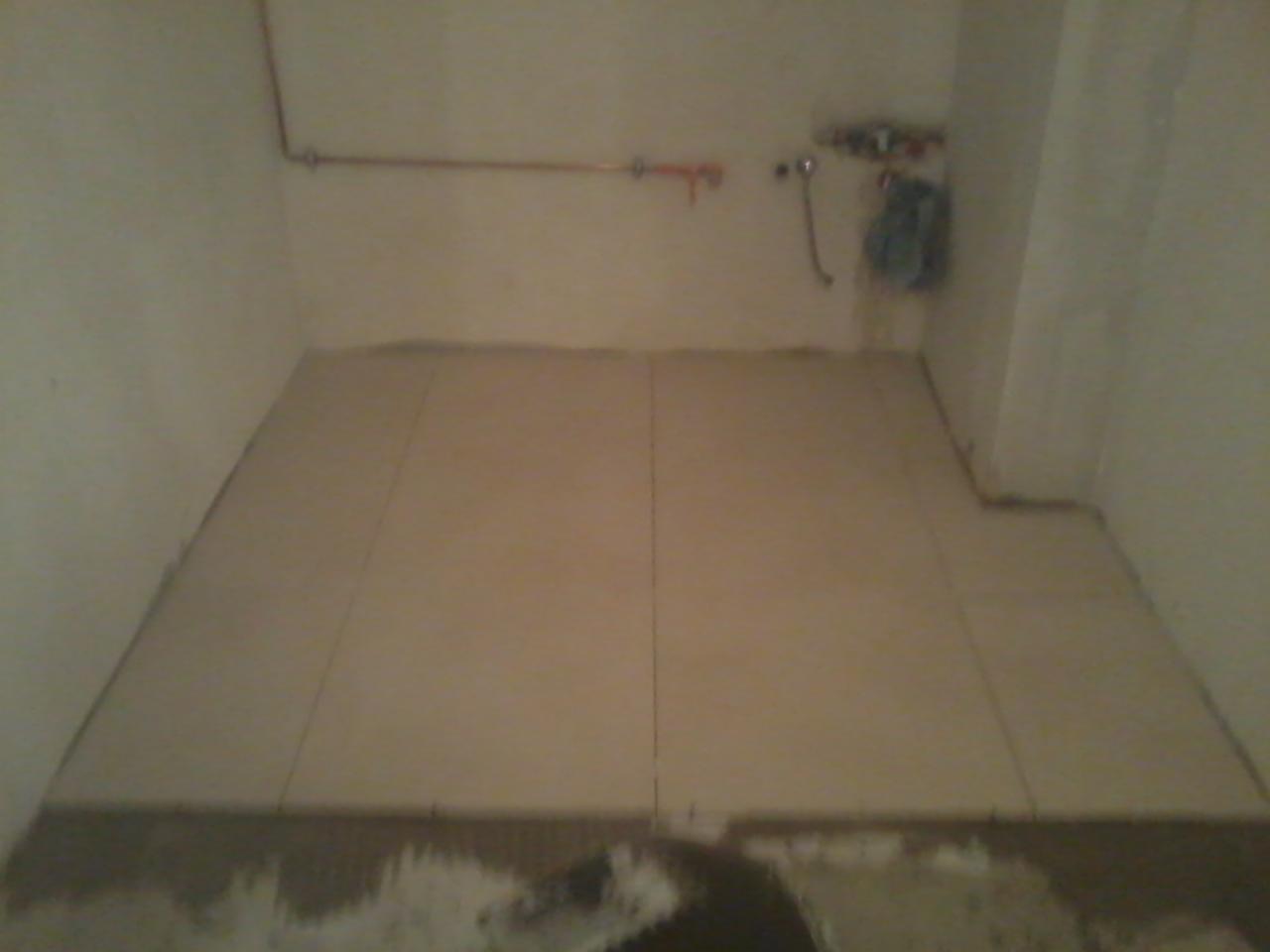 Ułożone, beżowe płytki podłogowe w trakcie prac w pomieszczeniu z instalacją wodną na ścianie. Widoczne fugi i niedokończone wykończenie przy podłodze.