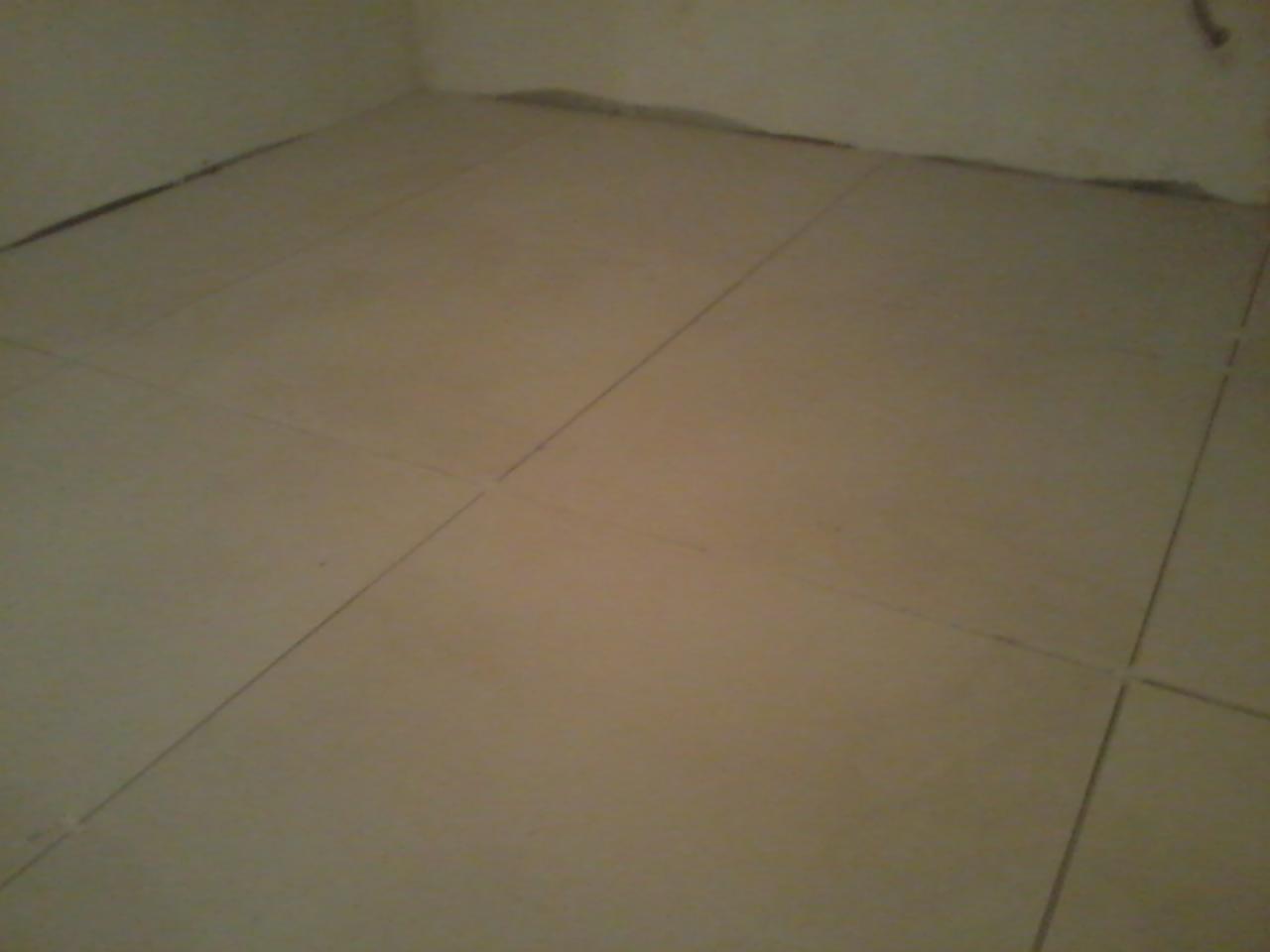 Beżowe płytki podłogowe ułożone w rogu pomieszczenia. Widoczne spoiny i niedokończone wykończenie przy ścianach. Brak listew przypodłogowych.