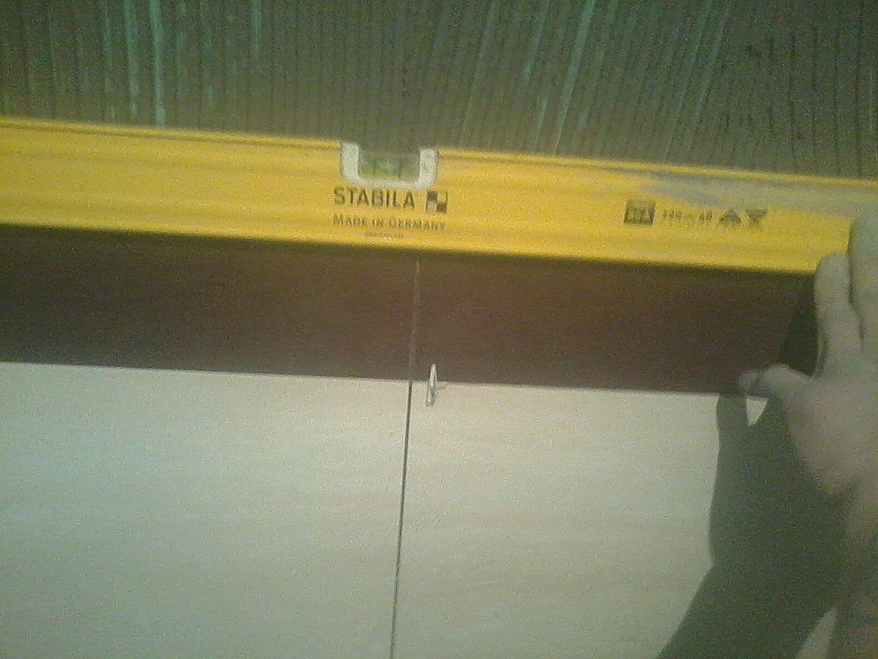 Żółta poziomica Stabila przyłożona do ściany, pion wyznacza linię, ręka stabilizuje narzędzie. Prace glazurnicze w trakcie. Ściana w kolorze złamanej bieli.