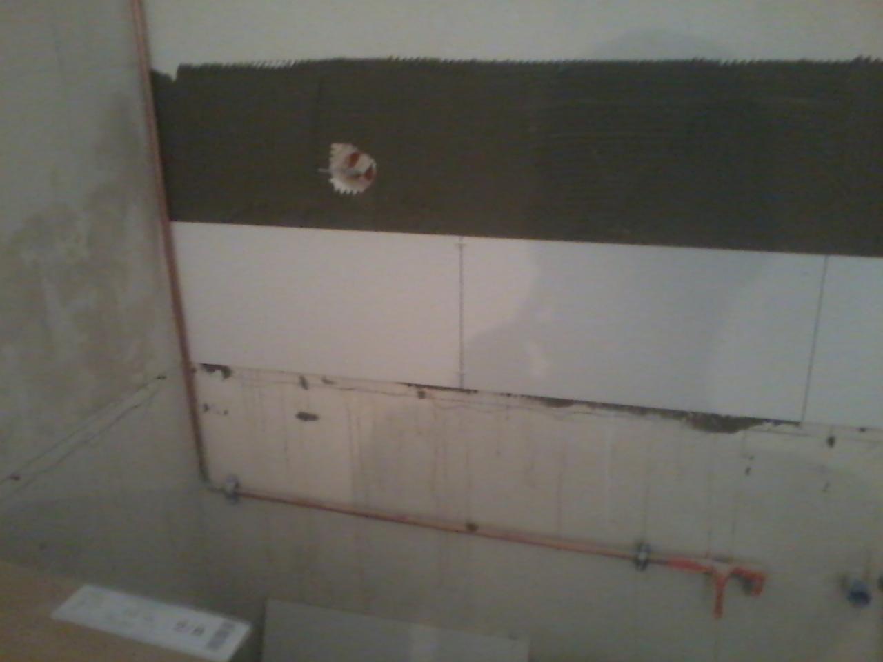 Układanie białych płytek na ścianie w łazience, widoczne ślady kleju i instalacja wodna. Prace glazurnicze w toku, surowy stan wykończenia.