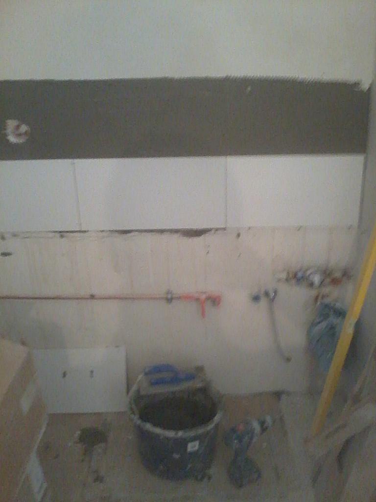 Ściana w trakcie układania płytek, widoczny pasek kleju i ułożone białe płytki. Na podłodze wiadro z zaprawą i narzędzia glazurnicze.