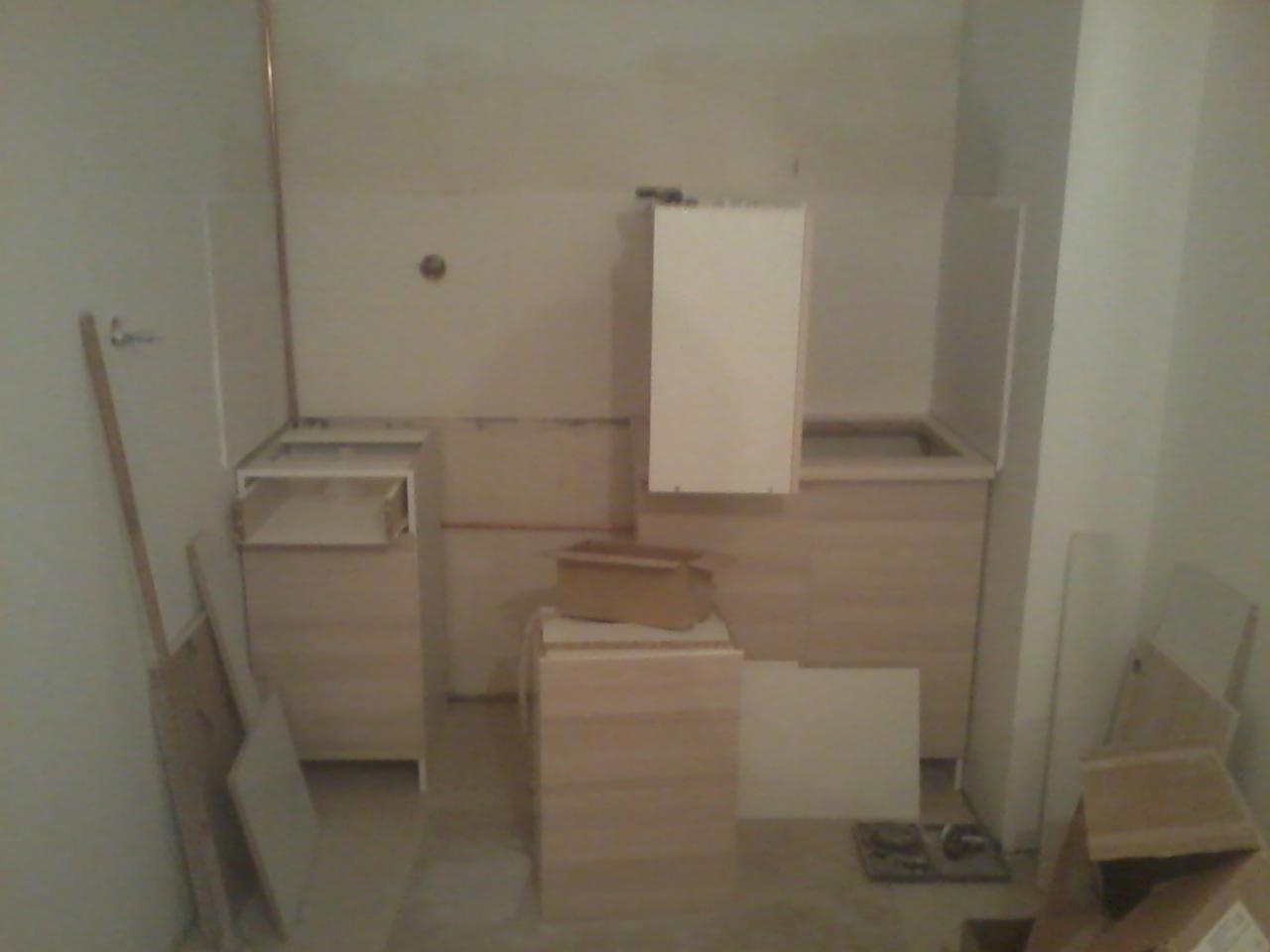 Łazienka w trakcie montażu mebli: szafki z jasnego drewna, biała wisząca szafka. Widoczne elementy instalacji i płyty. Prace wykończeniowe w toku.