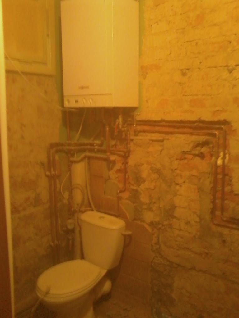 Łazienka w trakcie remontu w Dusznikach-Zdroju. Widoczne: toaleta, piec gazowy, odkryte rury miedziane i fragmenty starej ściany z cegły. Etap instalacji.