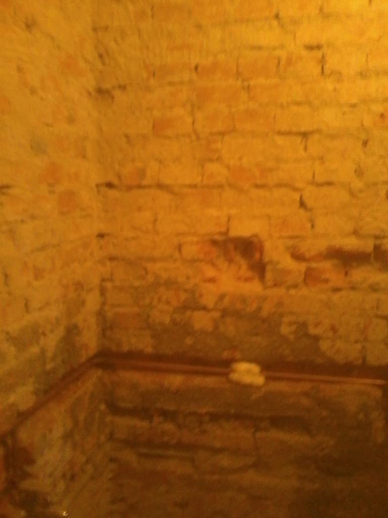 Narożnik z ceglaną, żółtawą ścianą w piwnicy z widoczną instalacją. Ściana wymaga renowacji, widoczne ubytki i stare fugi. Nierówna powierzchnia cegieł.