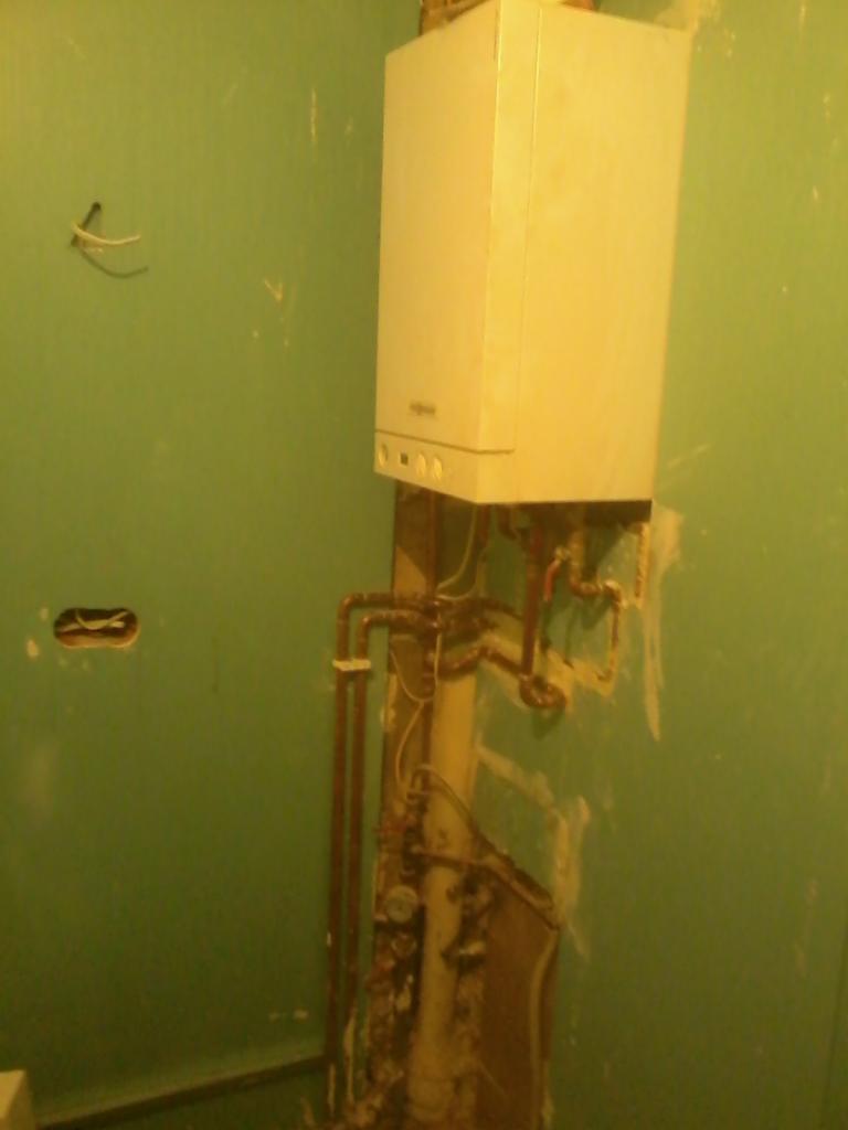 Bojler gazowy na zielonej ścianie w trakcie montażu, widoczne rury miedziane i ślady po remoncie. Ujęcie z bliska, widać niedoskonałości ściany i instalacji.