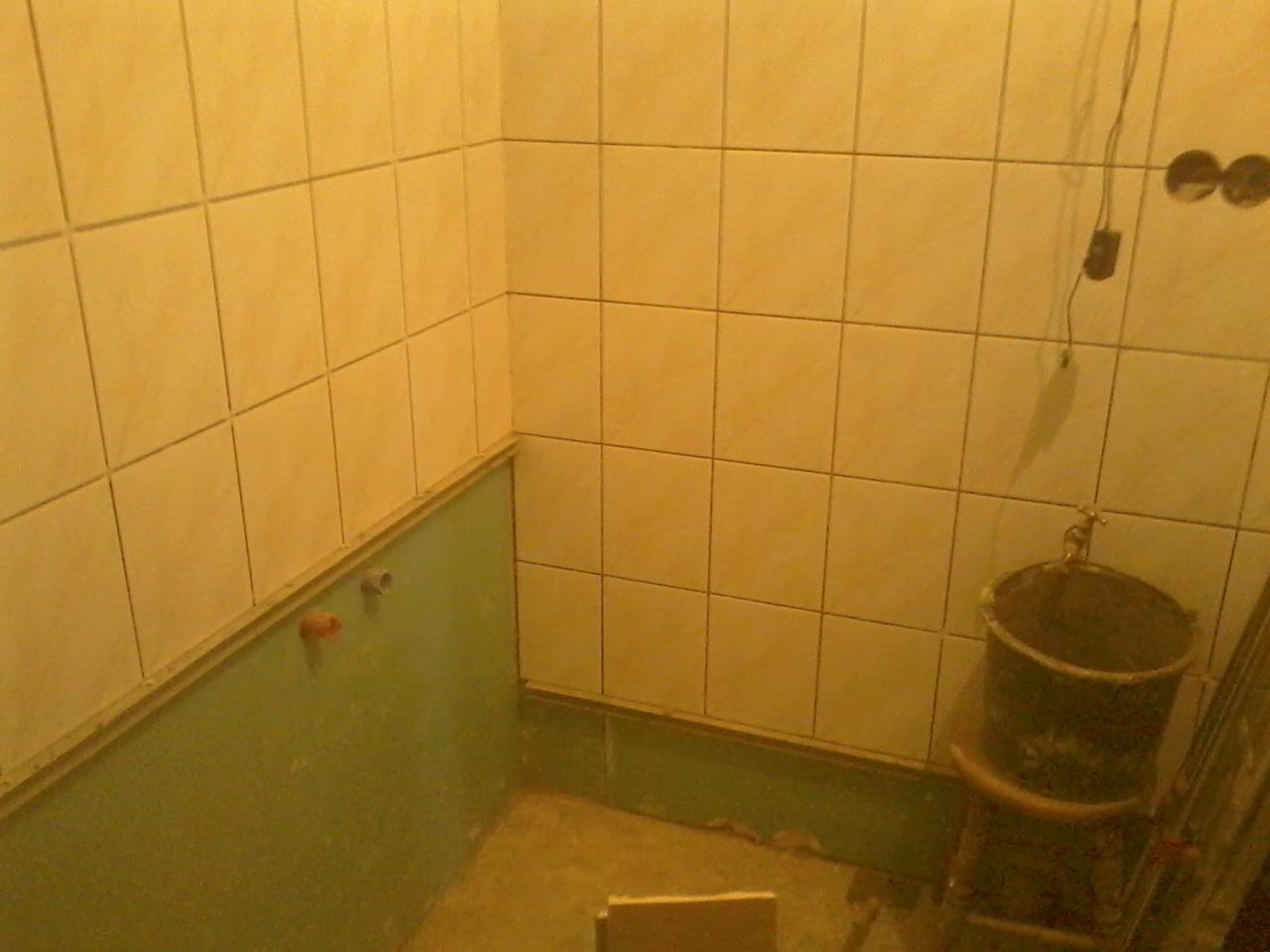 Ściany łazienki w trakcie remontu: częściowo położone kwadratowe, beżowe płytki, fragment instalacji wodnej. Widoczne też wiadro na stołku i kabel ze ściany.