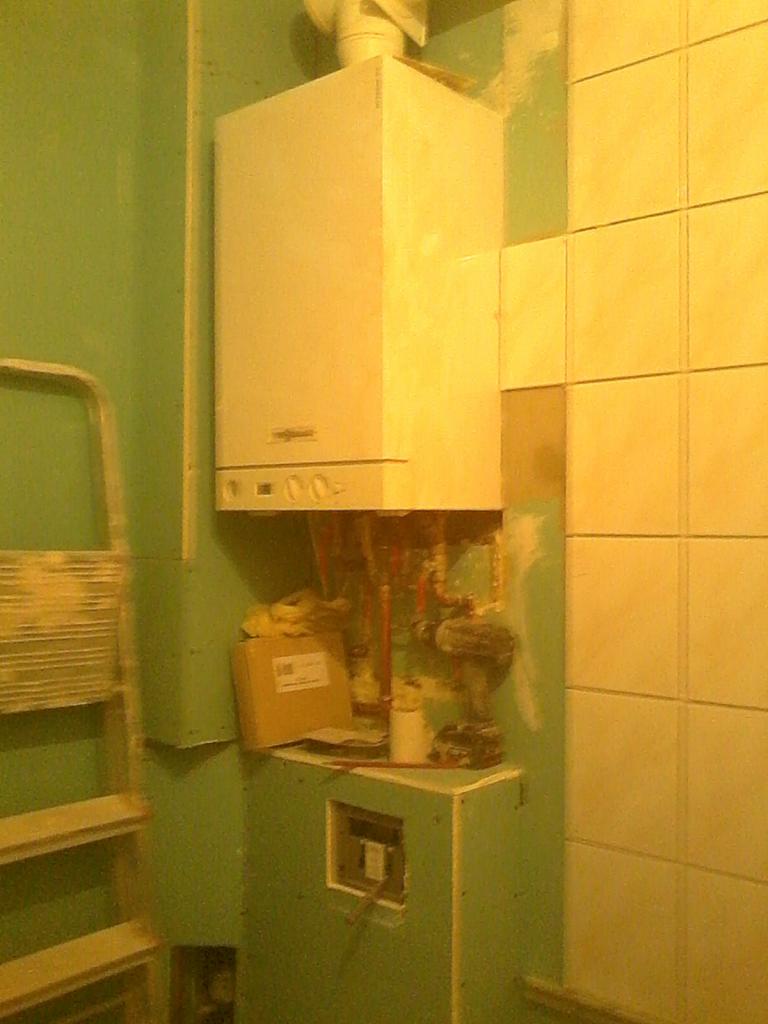 Montaż pieca gazowego w łazience z zielonymi ścianami i częściowo położonymi płytkami. Widoczna drabina i narzędzia.