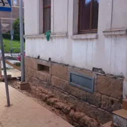 Odkopany kamienny fundament starego budynku z widocznymi śladami wilgoci, przygotowany do prac izolacyjnych, z fragmentem chodnika i ulicy w tle.