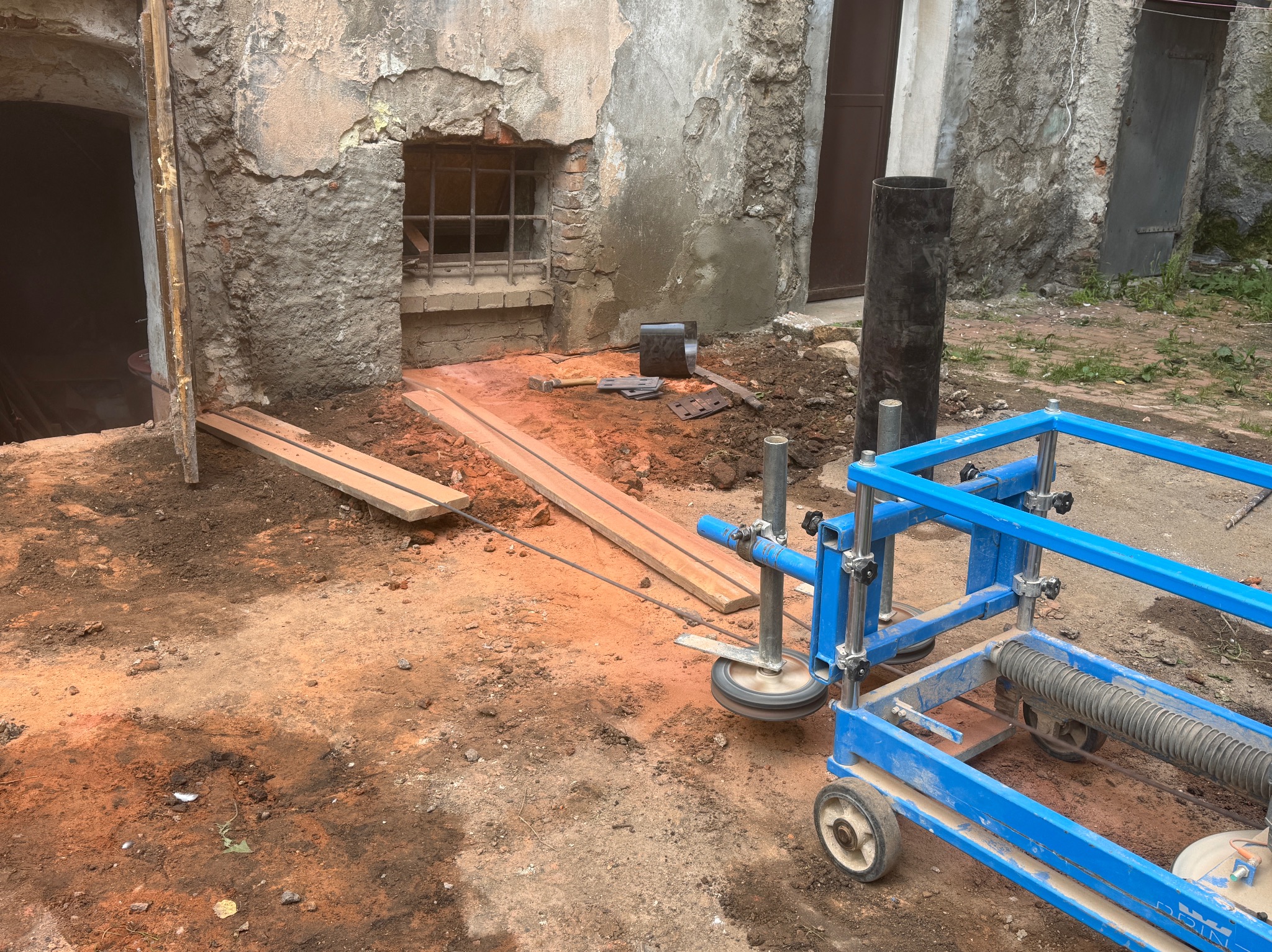 Przygotowanie do prac ziemnych przy fundamentach starego budynku z ceglanym oknem. Widoczny sprzęt budowlany i deski. Pomarańczowa ziemia.