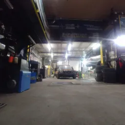 Wnętrze warsztatu samochodowego z widocznym samochodem bez maski w trakcie naprawy, maszyna do wyważania kół po lewej stronie, opony ułożone w stos po prawej stronie oraz baner reklamowy darmowego...