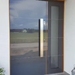 Nowoczesne, grafitowe drzwi wejściowe z długim, pionowym uchwytem ze stali nierdzewnej, odbijające krajobraz z domem w tle, zamontowane w jasnym budynku z szarą podłogą.