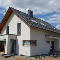 Nowoczesny dom jednorodzinny z jasną elewacją, szarą dachówką i roletami zewnętrznymi, widok z ogrodu w słoneczny dzień.