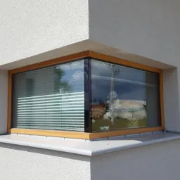 Narożne okno z drewnianą ramą, widoczne żaluzje wewnętrzne i odbicie otoczenia w szybie, elewacja budynku o jasnej, ziarnistej strukturze.