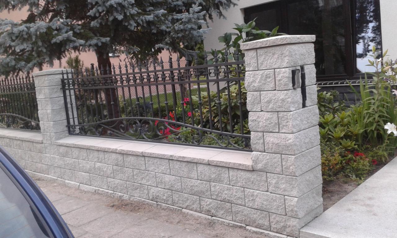 Kute przęsła ogrodzenia na podmurówce z szarej kostki, z ozdobnymi detalami i słupkami z tego samego materiału, widok na ogród z kwiatami i drzewami.