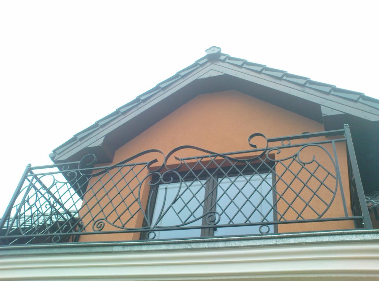 Kuta balustrada balkonowa z motywem rombów i spiralnych zakończeń, zamontowana na balkonie domu z pomarańczową elewacją i ciemnym dachem. Widok z dołu.