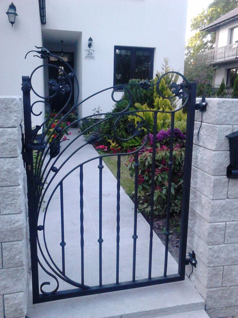 Kuta furtka z ozdobnymi elementami roślinnymi, zamontowana w kamiennym ogrodzeniu w Starej Łubiance, widok na ścieżkę i zadbany ogród.