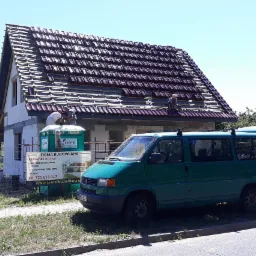 Budowa domu jednorodzinnego w trakcie kładzenia dachówki, widoczni dwaj dekarze na dachu, na pierwszym planie zielony bus firmy budowlanej i tablica informacyjna.